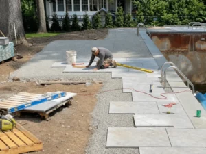 Worker laying large gray paving slabs on a gravel base beside an empty swimming pool.