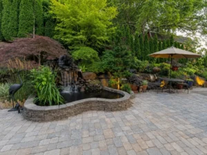 Garden paver patio beside a raised pond and waterfall, with an umbrella-shaded table and surrounding plants.