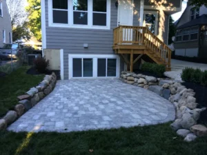 Curved paver patio bordered by large landscape rocks beneath a raised wooden entrance landing and stairs.