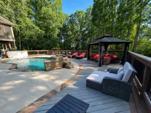 Poolside gray deck with a dark-roofed gazebo, red cushioned seating, and surrounding backyard trees.