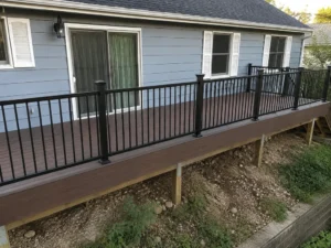 Long raised brown deck with black railings and exposed wooden support posts beside a gray house.