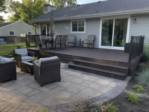 Dark brown deck with illuminated steps leading to a paver patio furnished with cushioned outdoor chairs.